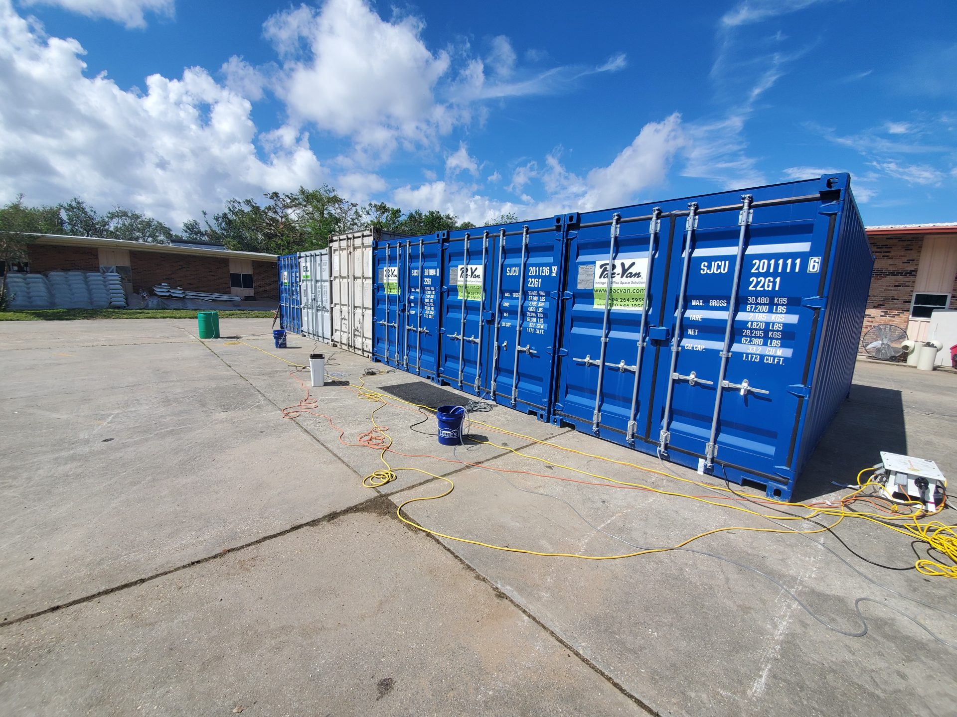 Blue shipping containers on a concrete lot under a sunny sky, with cables and tools nearby