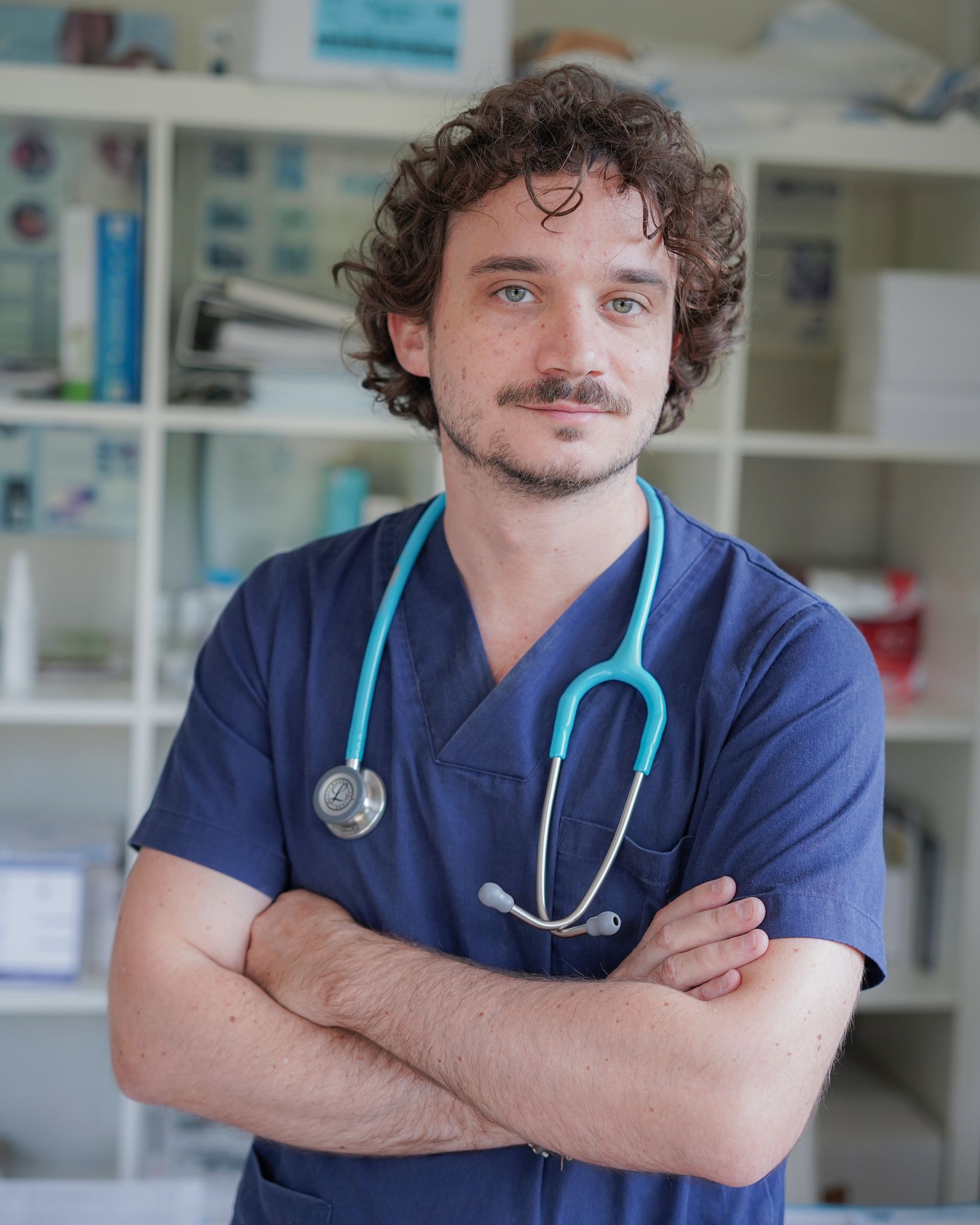 Uomo con i capelli ricci, che indossa un camice blu e uno stetoscopio, con le braccia incrociate, in uno studio medico.