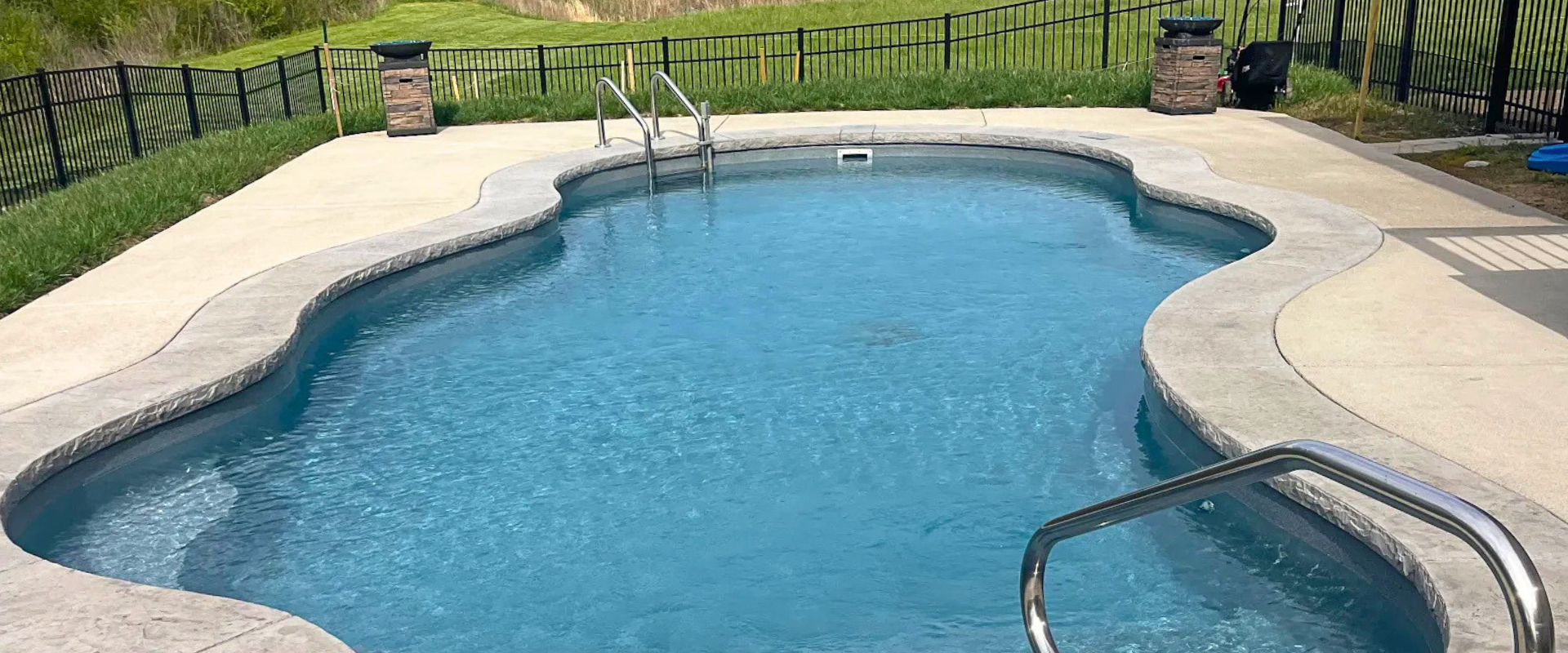 Backyard swimming pool with clear blue water, metal handrails, and a fenced lawn in the background