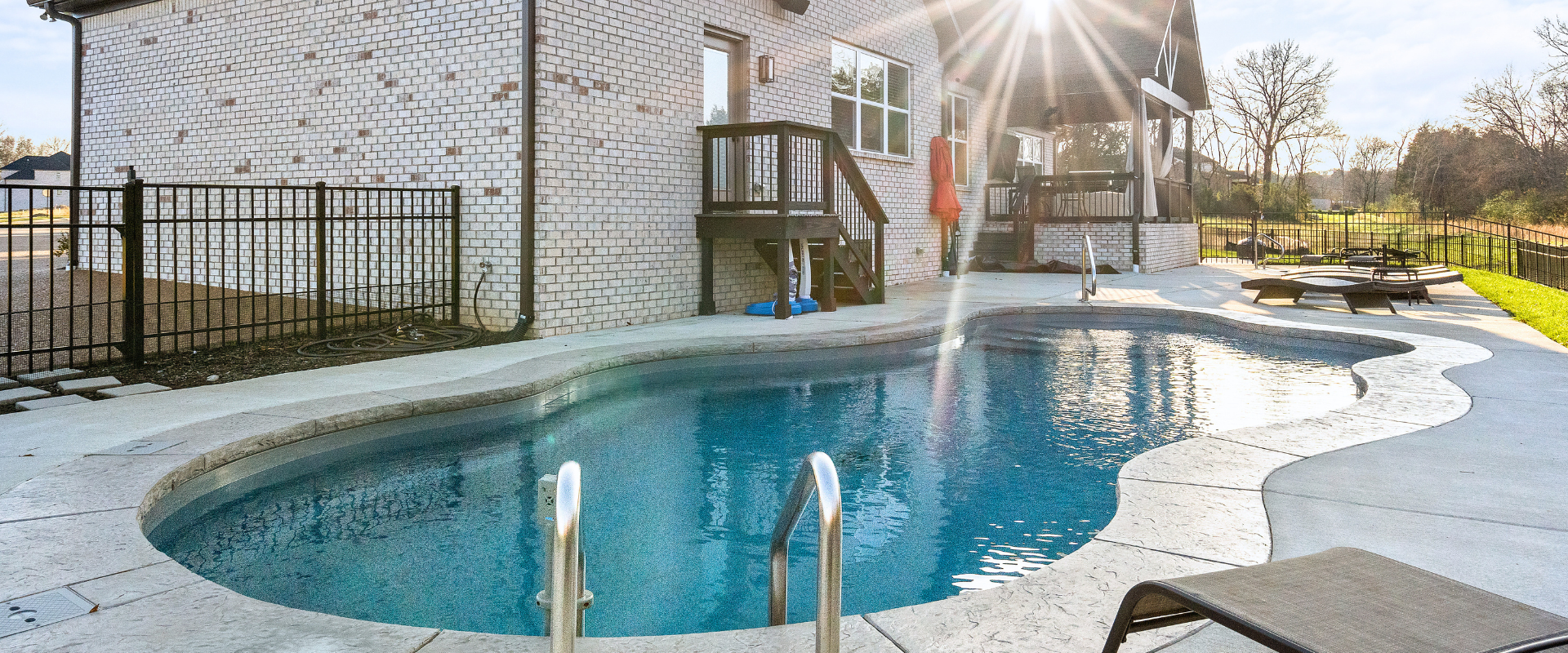 Backyard pool with curved blue water, patio chairs, and bright sunlight near a white house