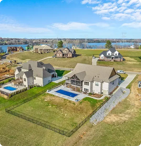 Aerial view of suburban houses with fenced yards and blue pools near a waterfront under a bright sky