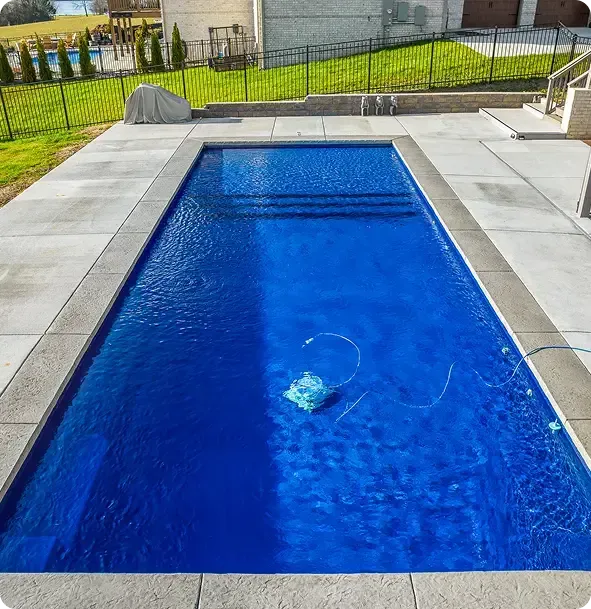 Rectangular blue swimming pool with a pool cleaner, bordered by concrete patio and a fenced yard