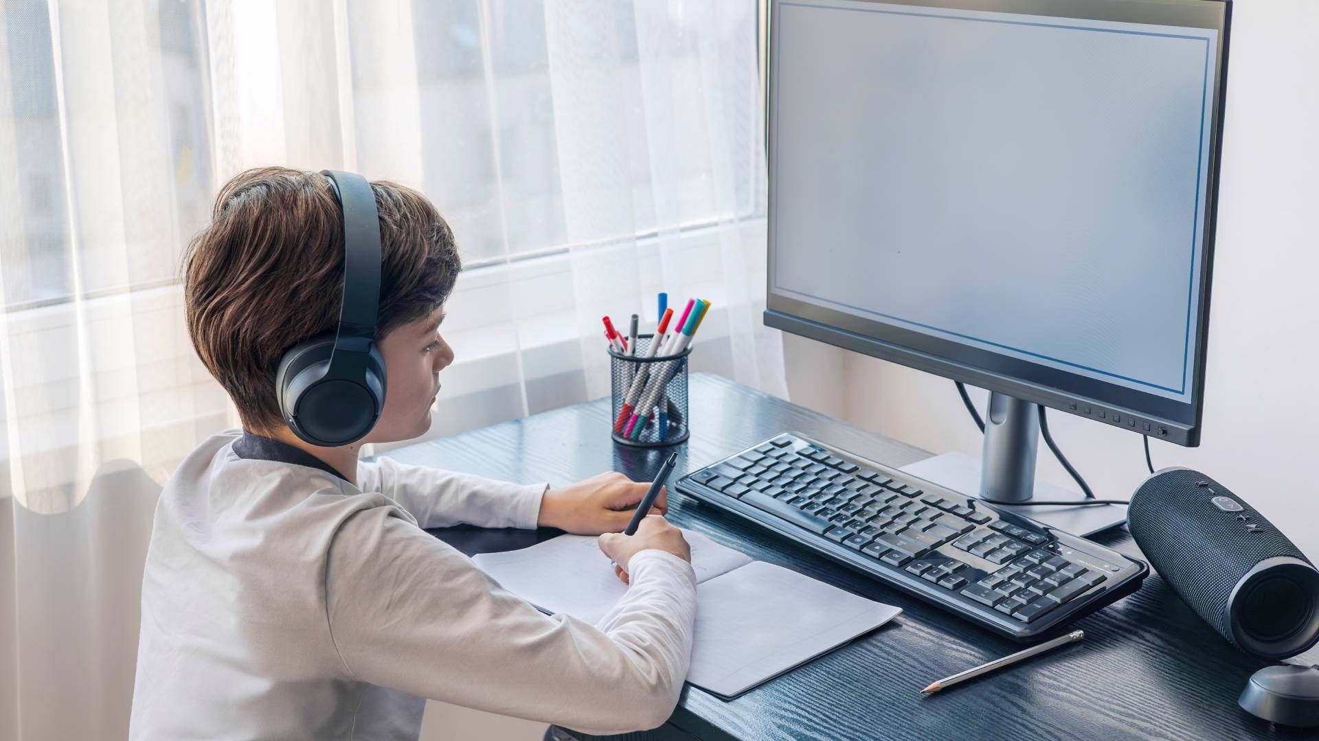 Boy with headphones, writing at a desk, looking at a computer monitor.