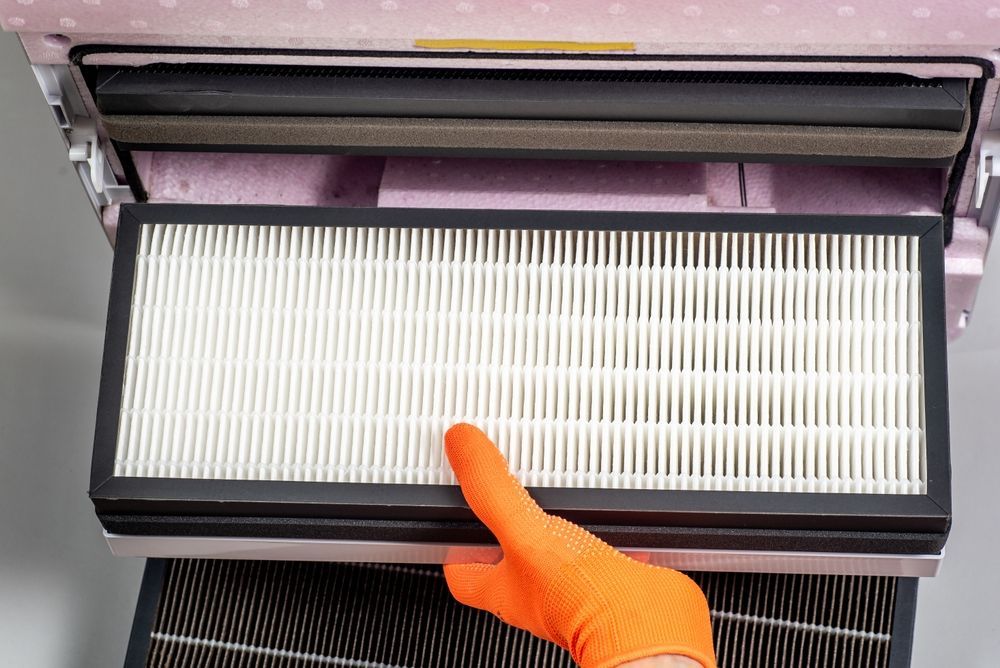 Gloved hand pointing at a white HVAC filter inside an open vent compartment