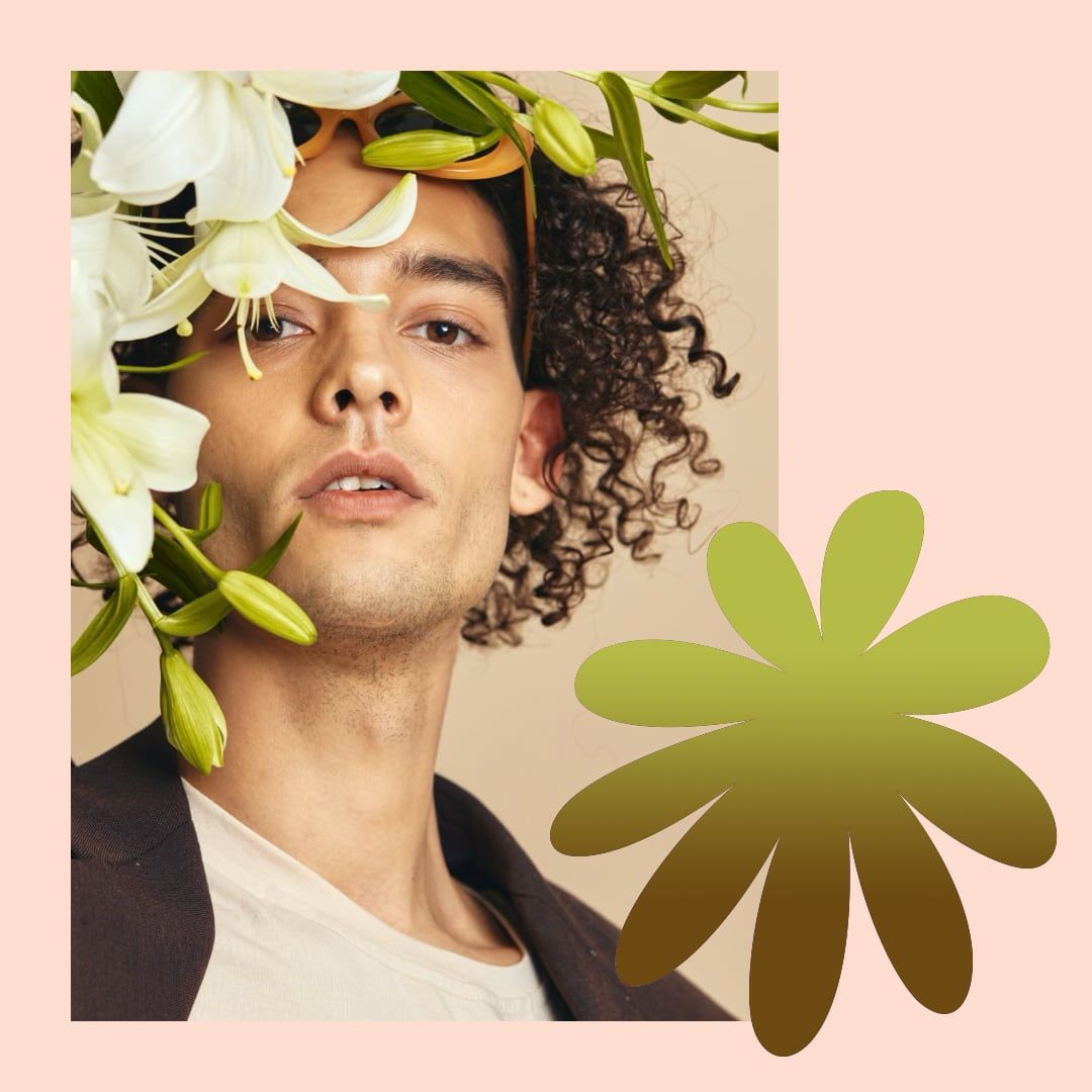 Portrait of a person with curly hair beside white flowers and green leaves on a pink background