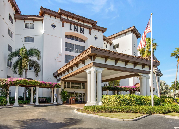 Exterior of a white hotel with a covered entrance, palm trees, and flags under a blue sky