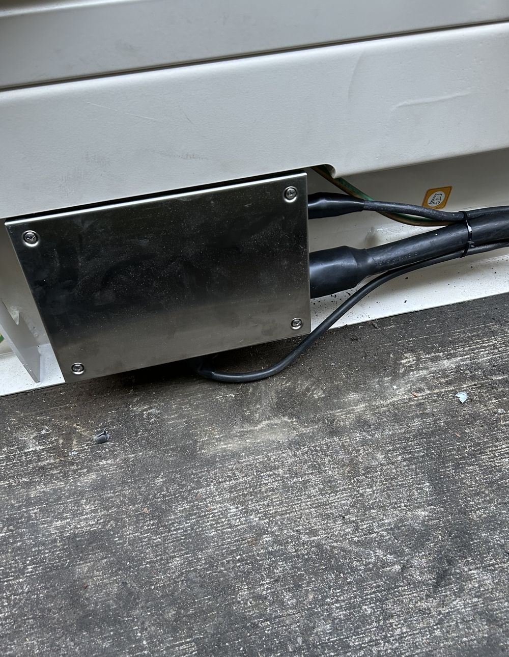 A metal box bolted to a white surface, with black cables extending out, resting on a gray floor.