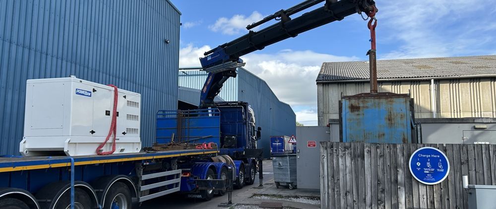 A crane lifting an object into a metal bin near a large truck and industrial buildings.