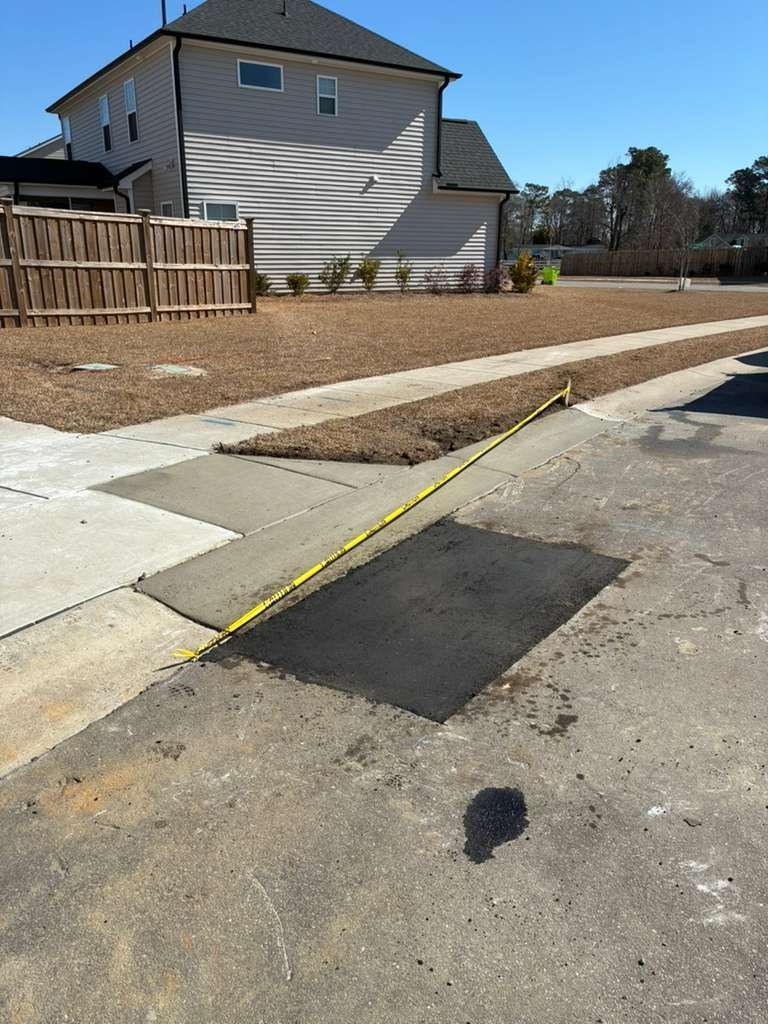 Sidewalk ramp and curb cut beside a residential street, with a yellow caution line and fresh patchwork pavement.