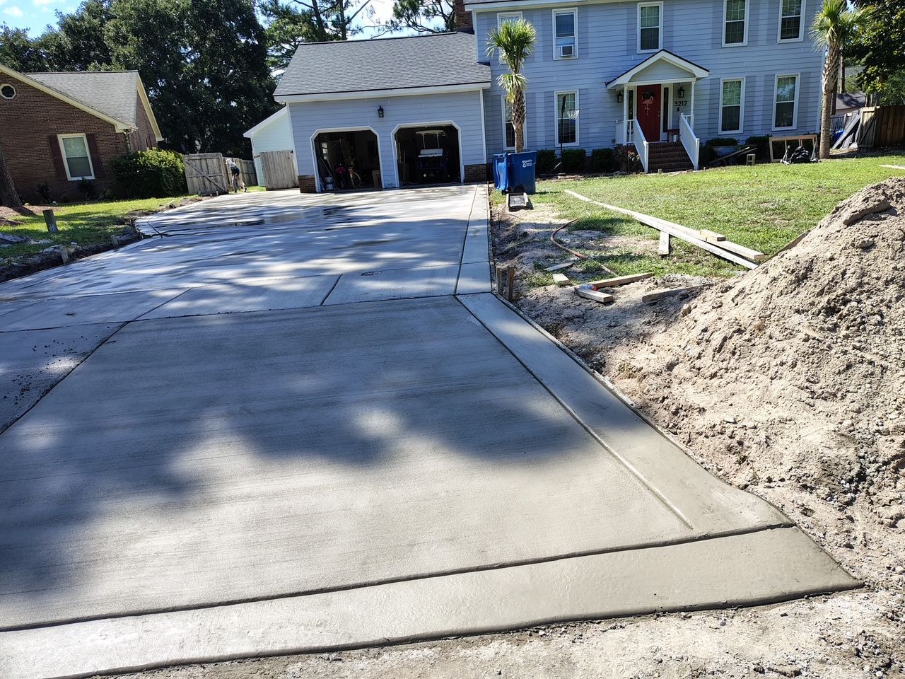 Concrete driveway under construction beside a house, with soil piles and a garage in the background.