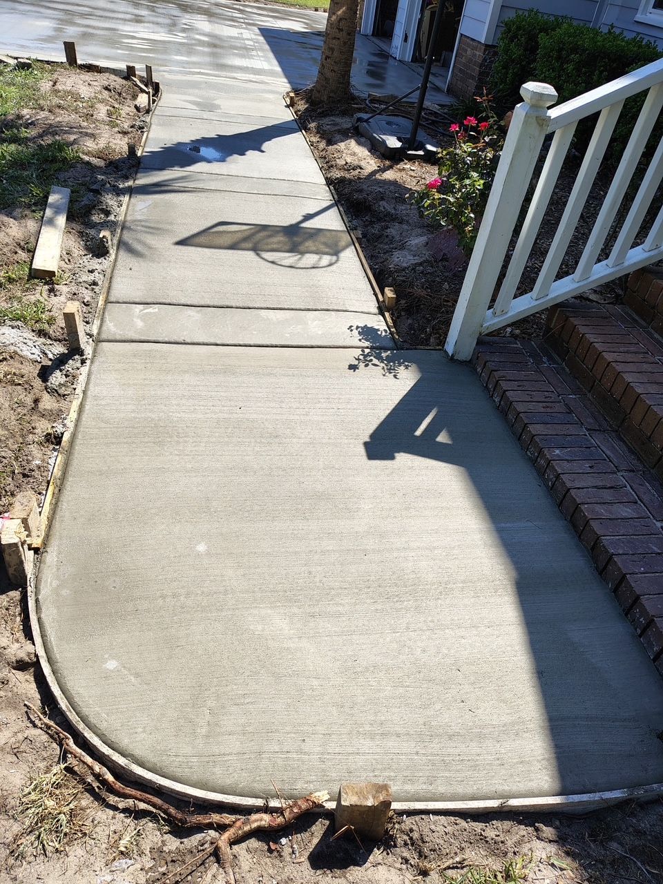 Broken concrete walkway with a cracked edge beside a white porch railing and garden bed.
