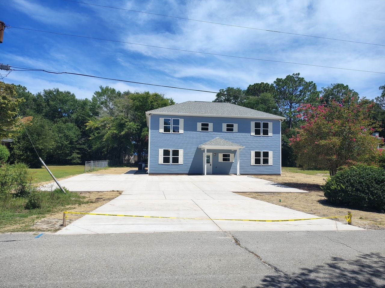 Two-story light blue house with white trim and a driveway in front, surrounded by trees under a blue sky