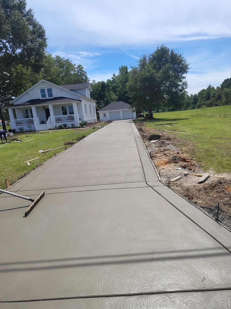 Long concrete driveway beside a white house and garage, with grassy land and trees under a blue sky.