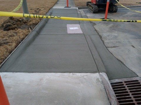 Freshly poured concrete sidewalk ramp with caution tape across it and a storm drain grate nearby