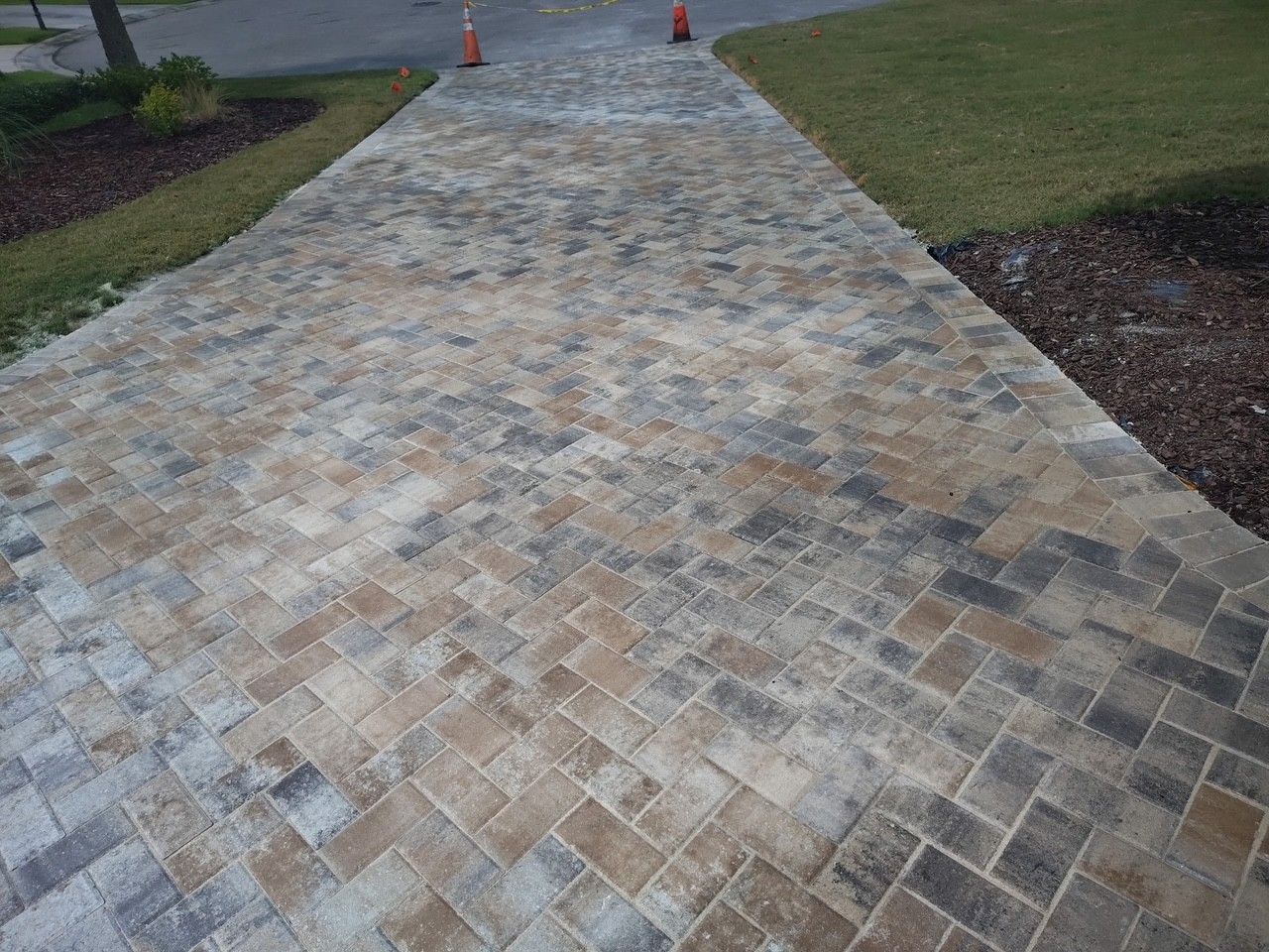 Brick paver walkway leading to a road, bordered by grass and mulch, with orange cones ahead
