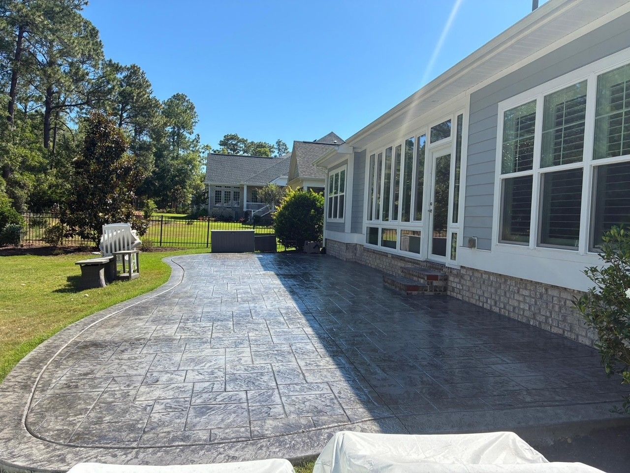 Stamped concrete patio beside a white house with large windows, lawn and trees in bright sunlight