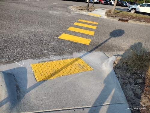 Street curb ramp with yellow tactile paving leading to painted crosswalk stripes in a parking lot area