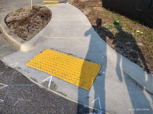 Sidewalk curb ramp with yellow tactile paving at a concrete path intersection beside a road and landscaped area