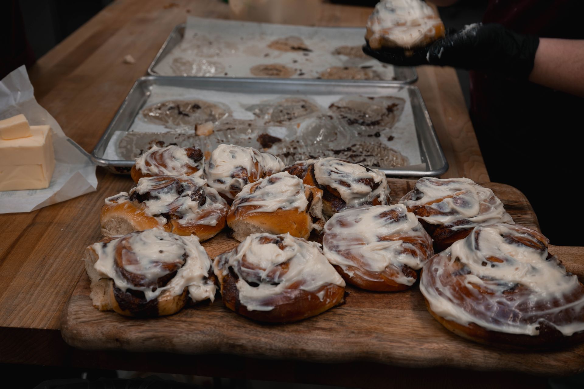 Tray of cinnamon rolls with white icing on a wooden table, with a hand serving one roll