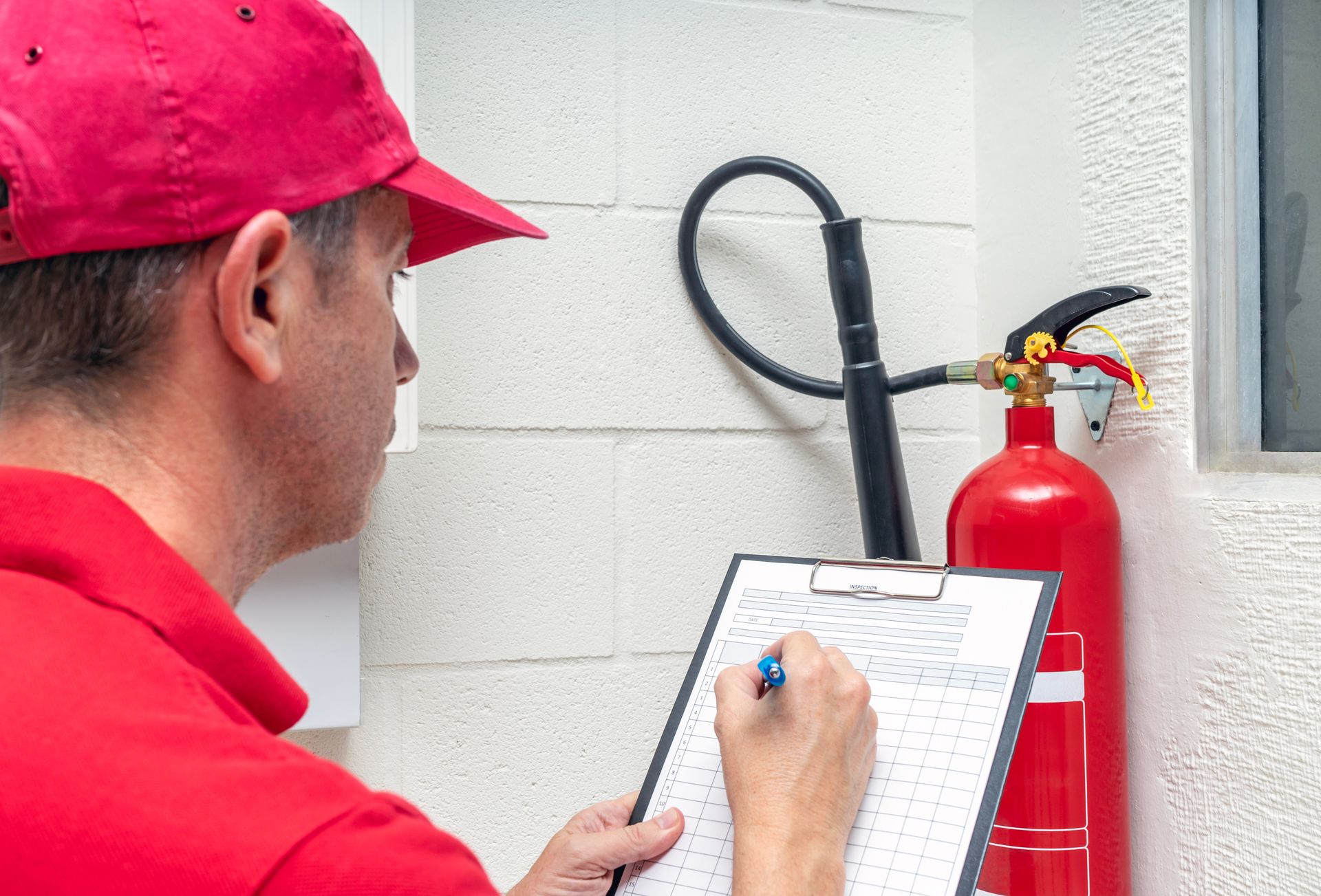 A man in a red hat is writing on a clipboard next to a fire extinguisher.