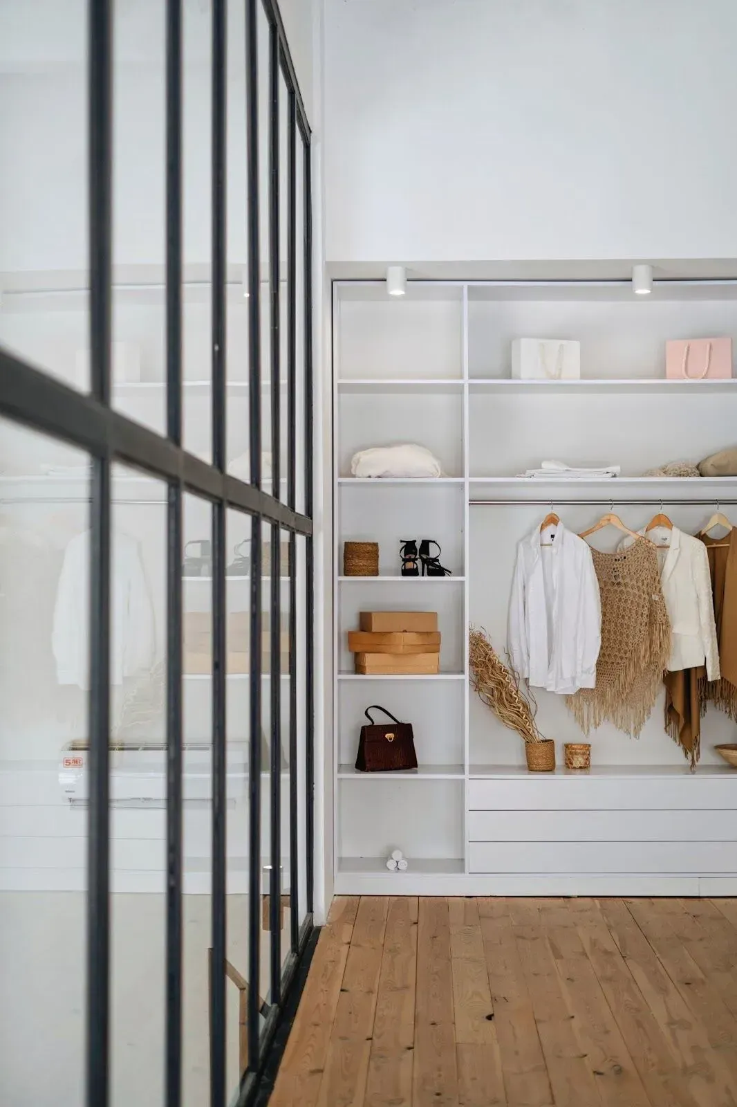 Modern walk-in closet with white open shelving, hanging clothes, and light wood flooring, viewed behind a black grid wall.