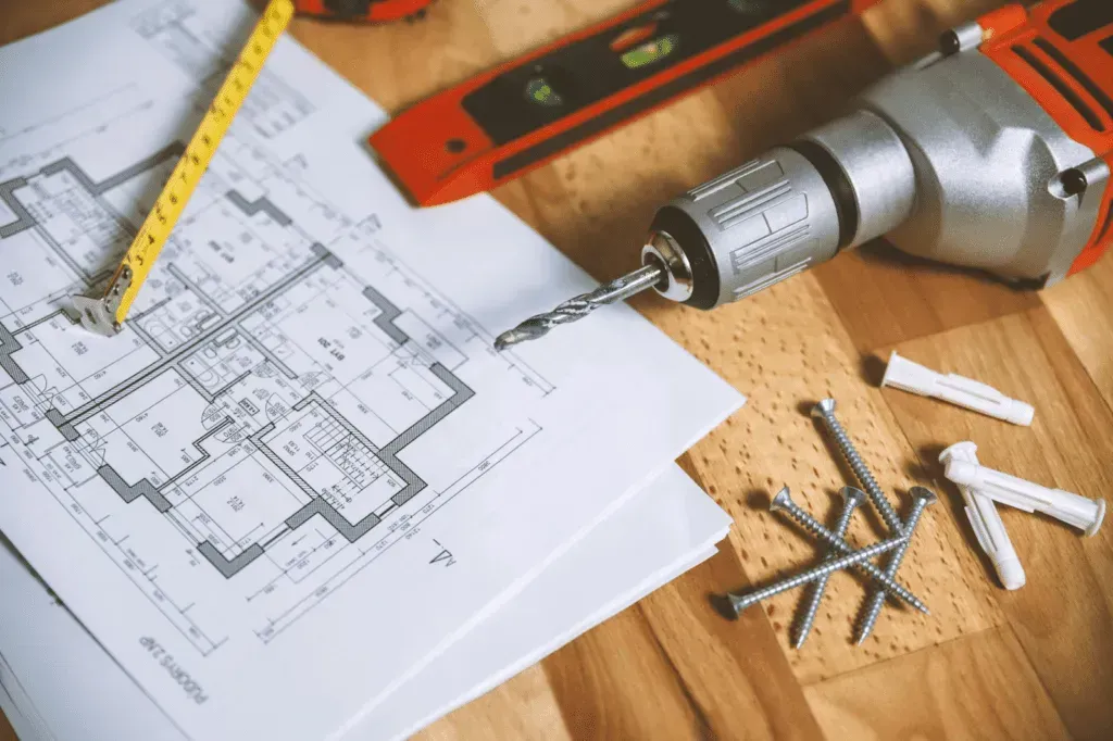 A floor plan, tape measure, level, drill, and screws laid out on a wooden surface for a home renovation project.