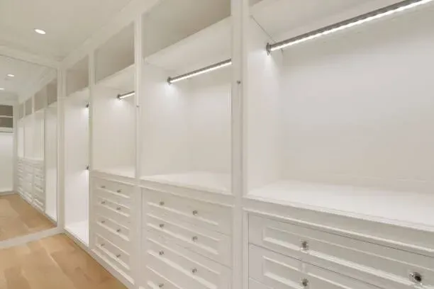 Modern walk-in closet with white shelving, built-in drawers, illuminated clothing rods, and light hardwood flooring.