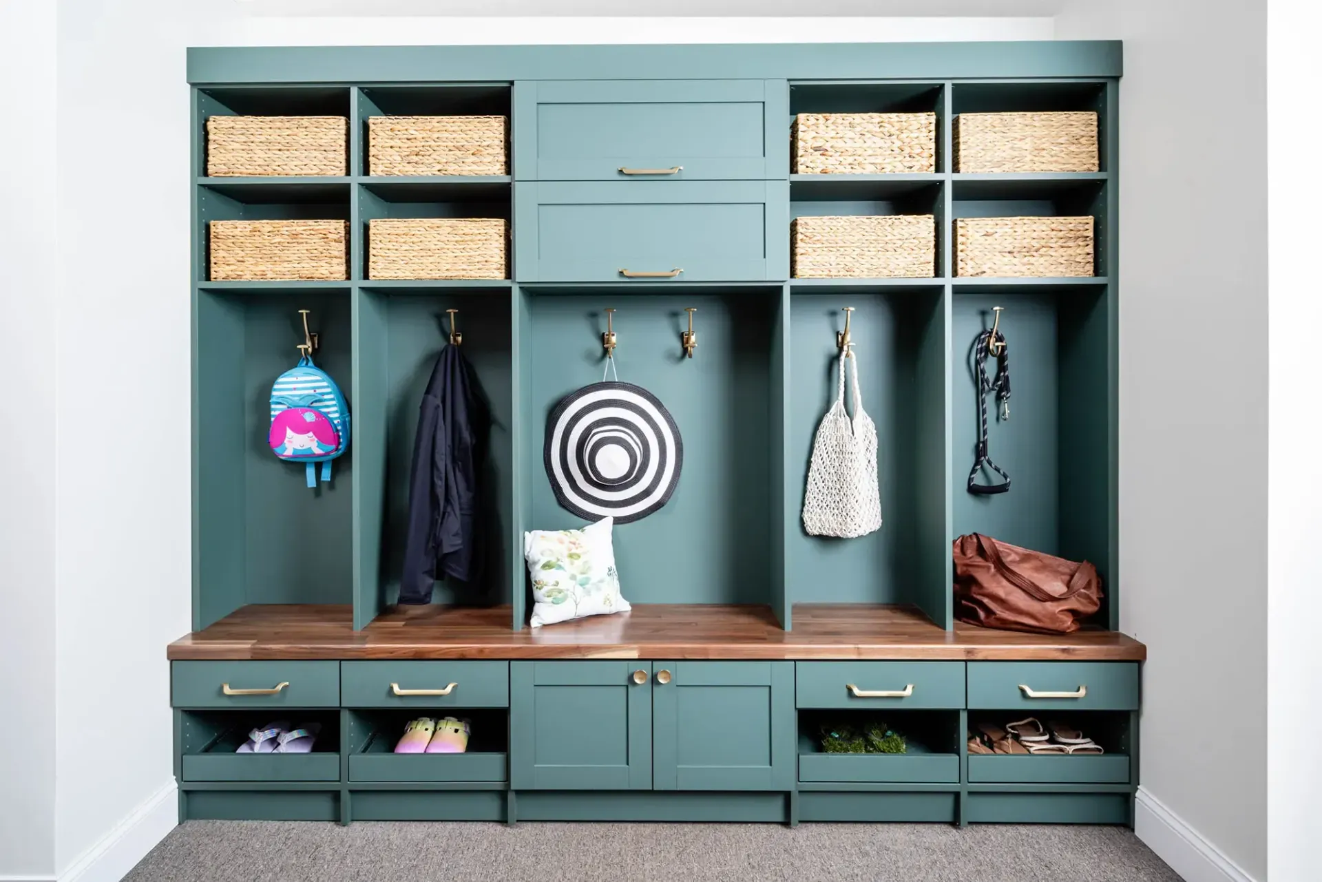 A dark teal mudroom built-in unit with storage cubbies, hooks for hanging items, upper baskets, and lower shoe drawers.