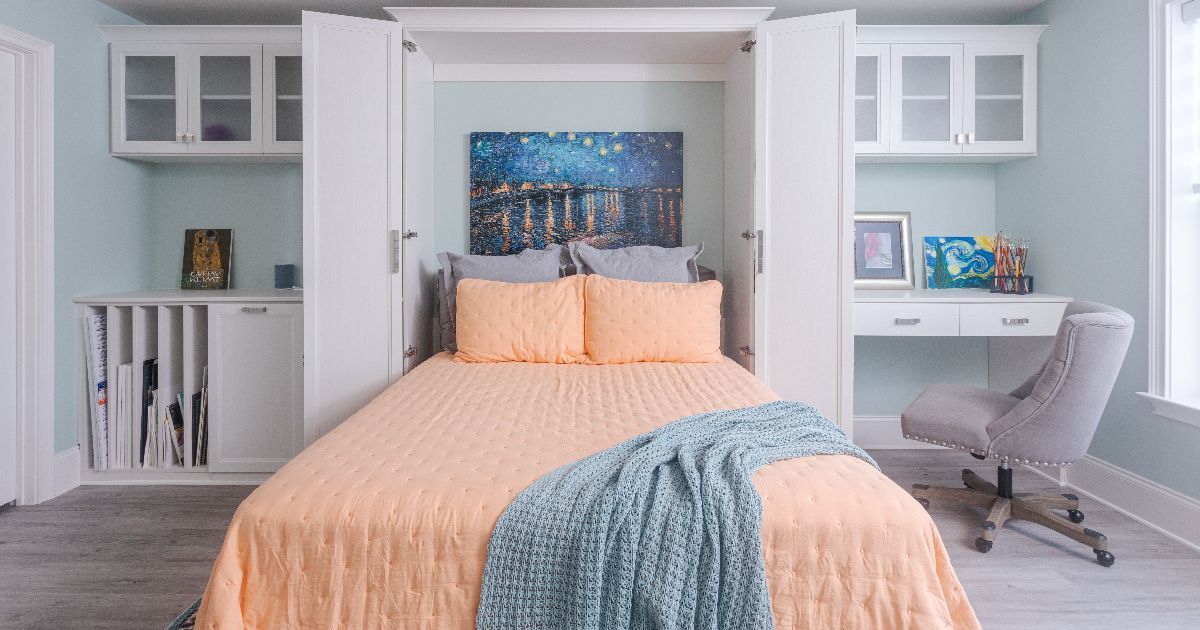 A murphy bed with peach bedding and a blue throw sits between white storage cabinets and a desk with a grey chair.
