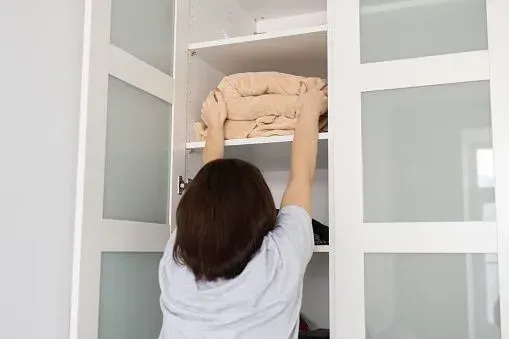 A person stands reaching up into a white cabinet to place or retrieve a folded beige blanket from a high shelf.