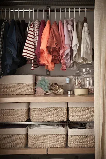 A closet with clothes hanging on a rack above two shelves containing multiple woven storage bins and glass jars.