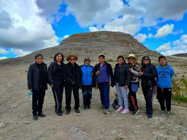 Grupo de personas de pie sobre terreno rocoso con una colina escalonada y un cielo azul nublado detrás.