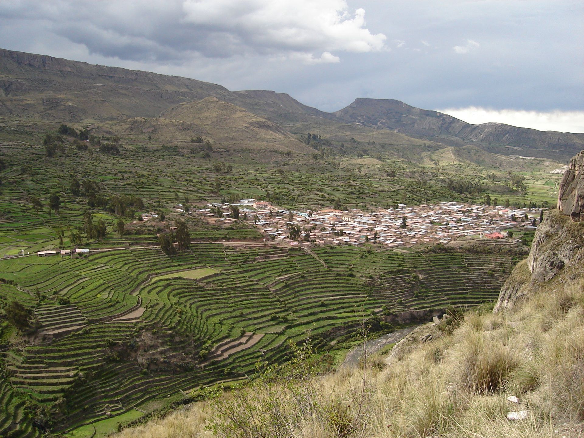 Laderas verdes aterrazadas rodean un pequeño pueblo en un valle de montaña bajo cielos nublados.