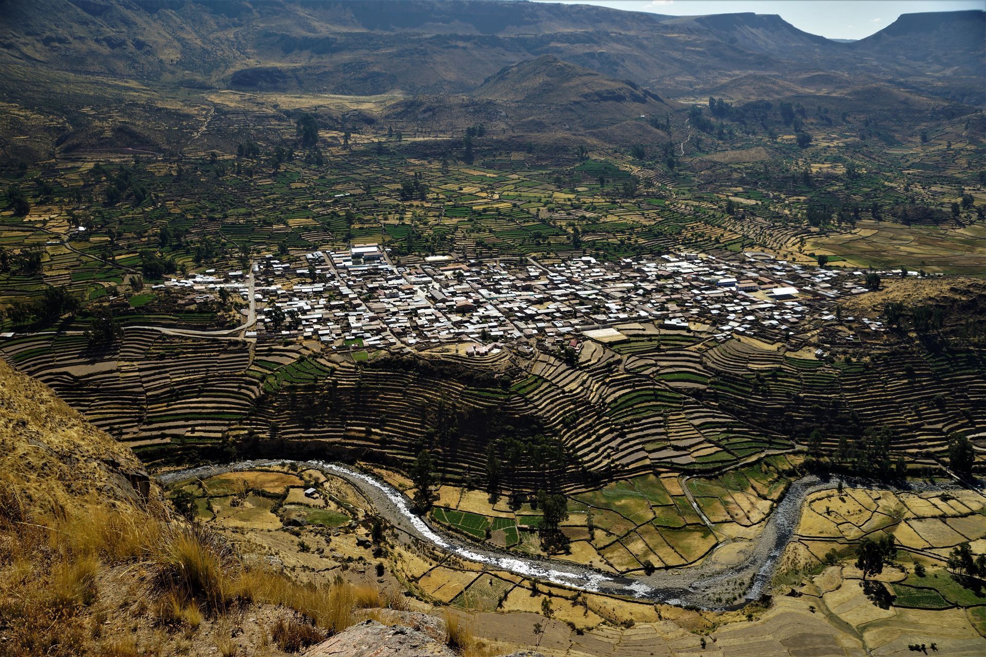 Pueblo de valle de montaña con campos en terrazas, río serpenteante y verdes colinas bajo un cielo nublado.