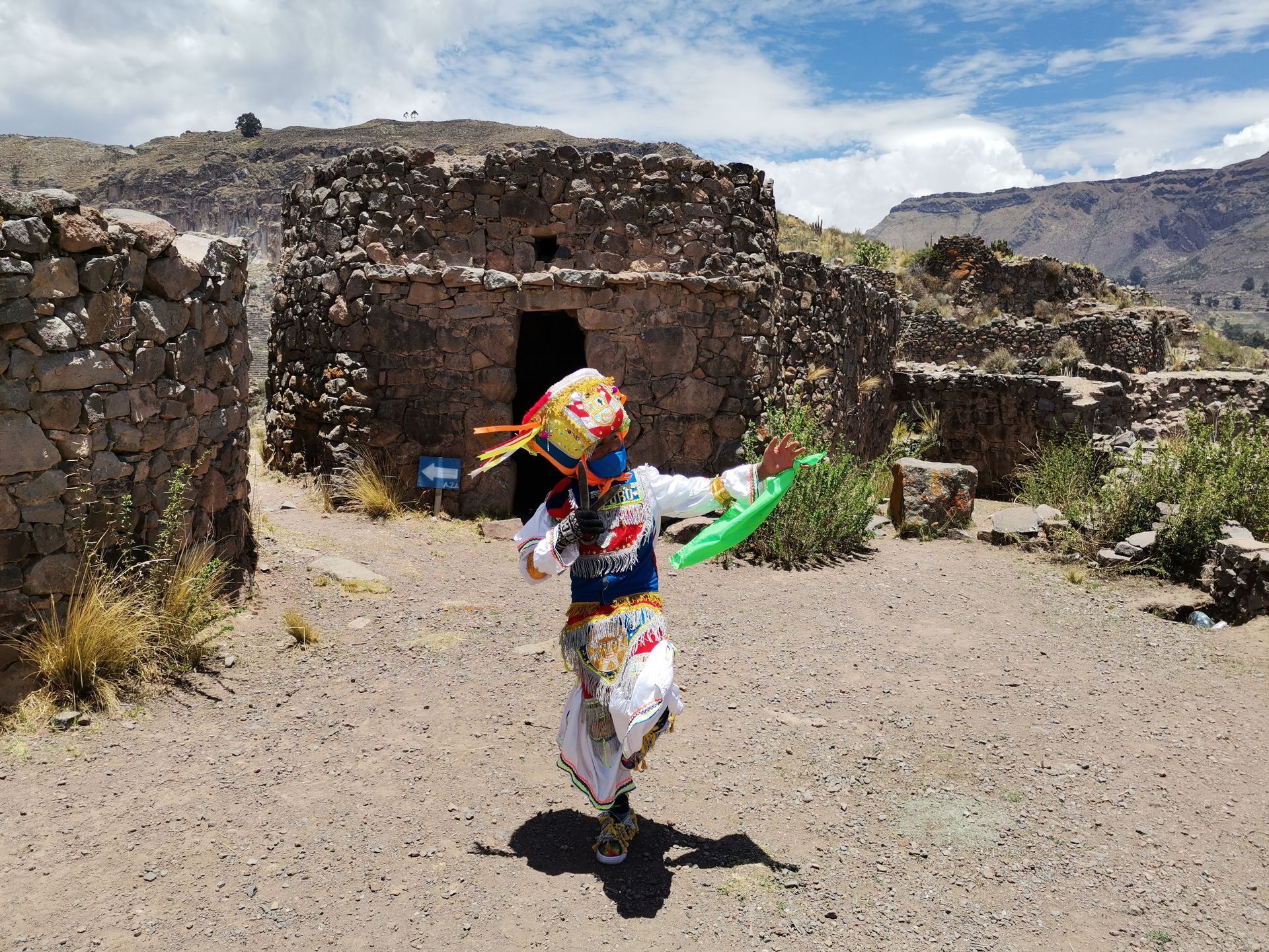 Una persona con un traje colorido baila al aire libre frente a un edificio de piedra en un sendero rocoso.