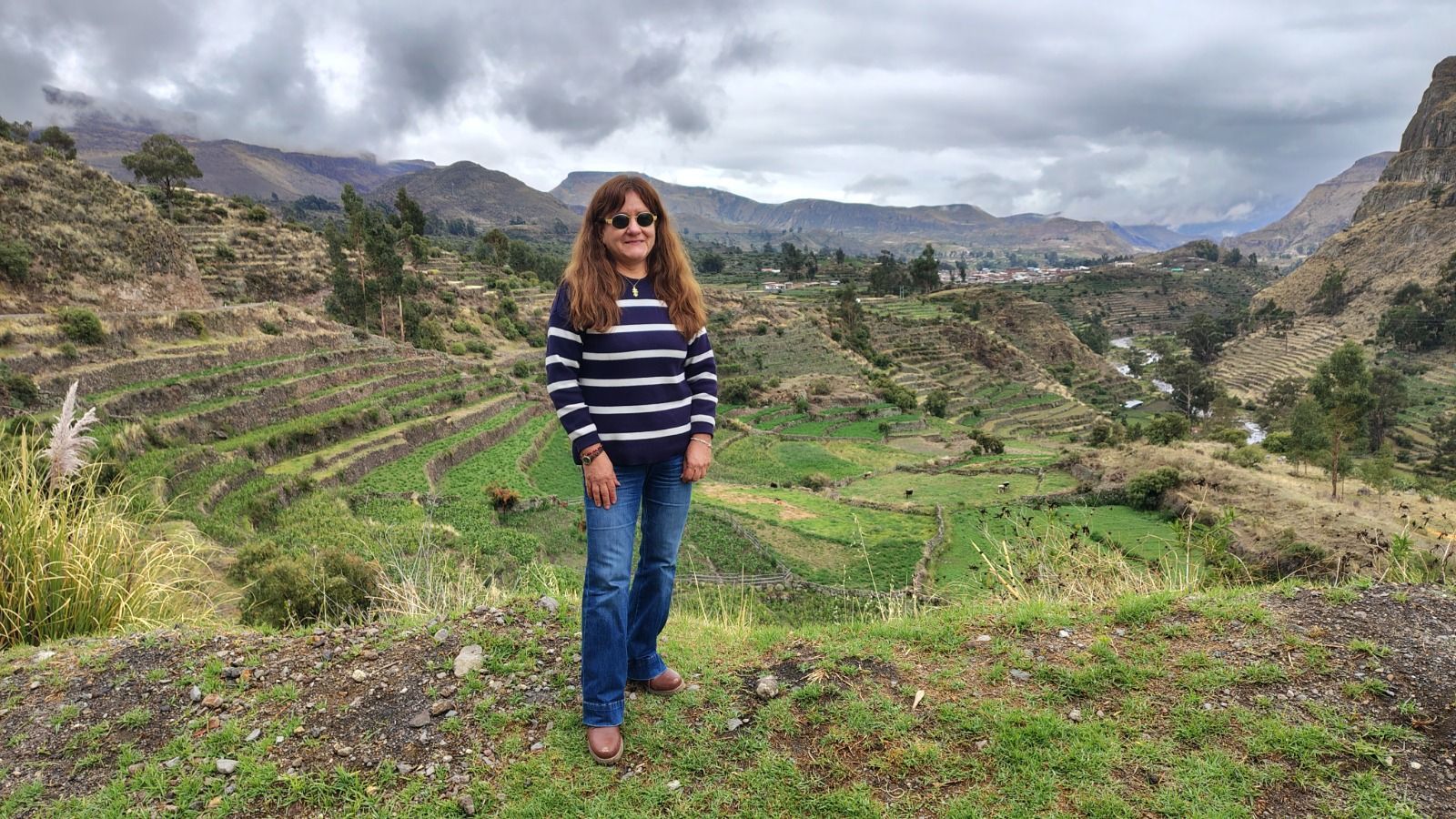 Mujer de pie en una ladera verde aterrazada bajo montañas nubosas, vestida con un suéter de rayas y jeans.