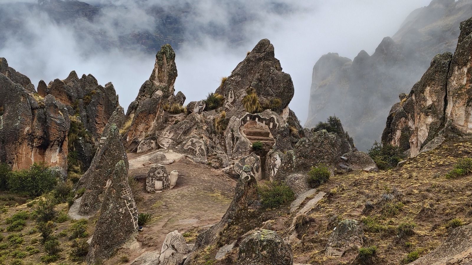 Sendero de montaña rocoso con altas cumbres escarpadas y nubes brumosas al fondo.