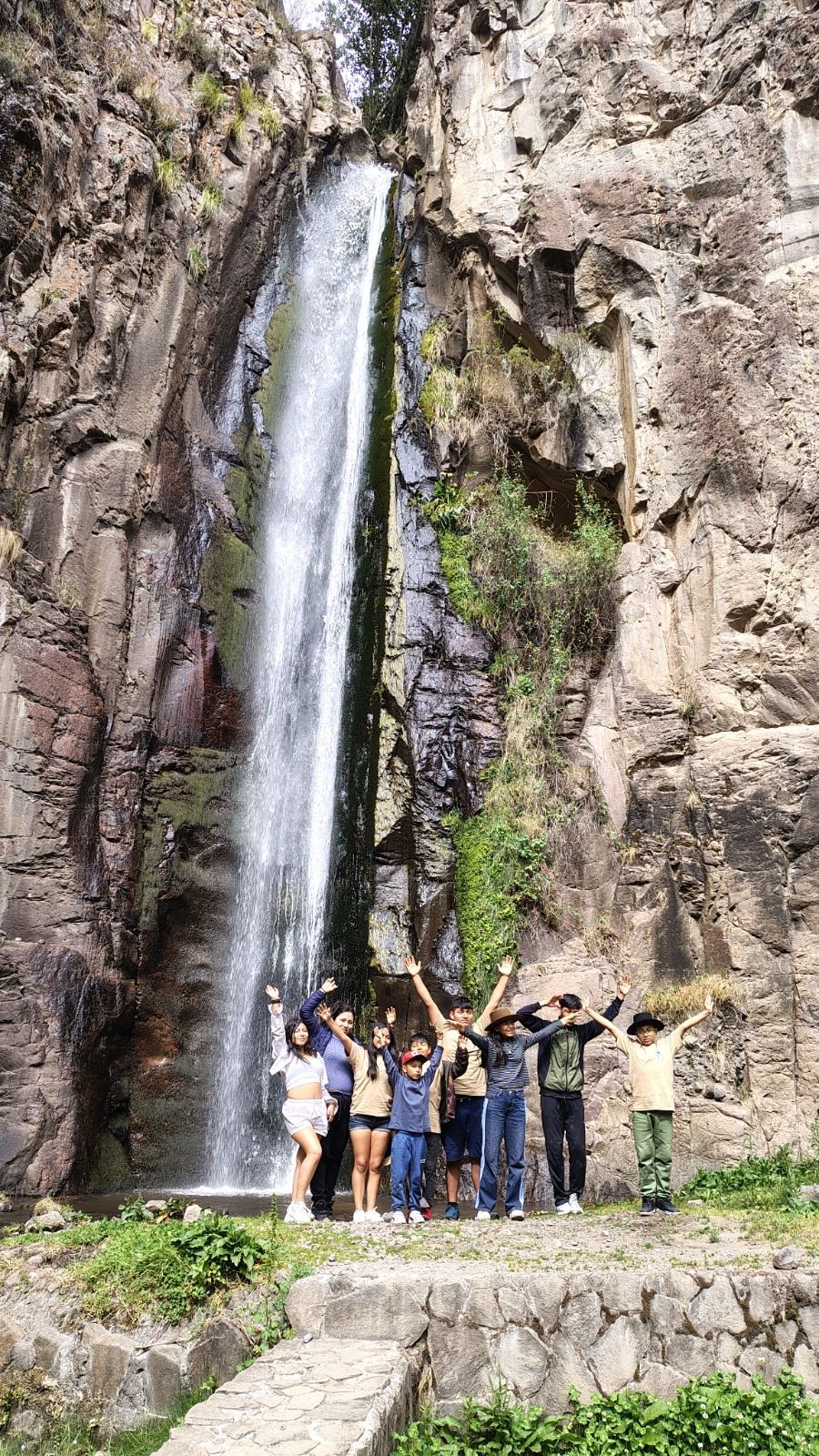 Personas posando al pie de una alta cascada, junto a acantilados rocosos y vegetación.