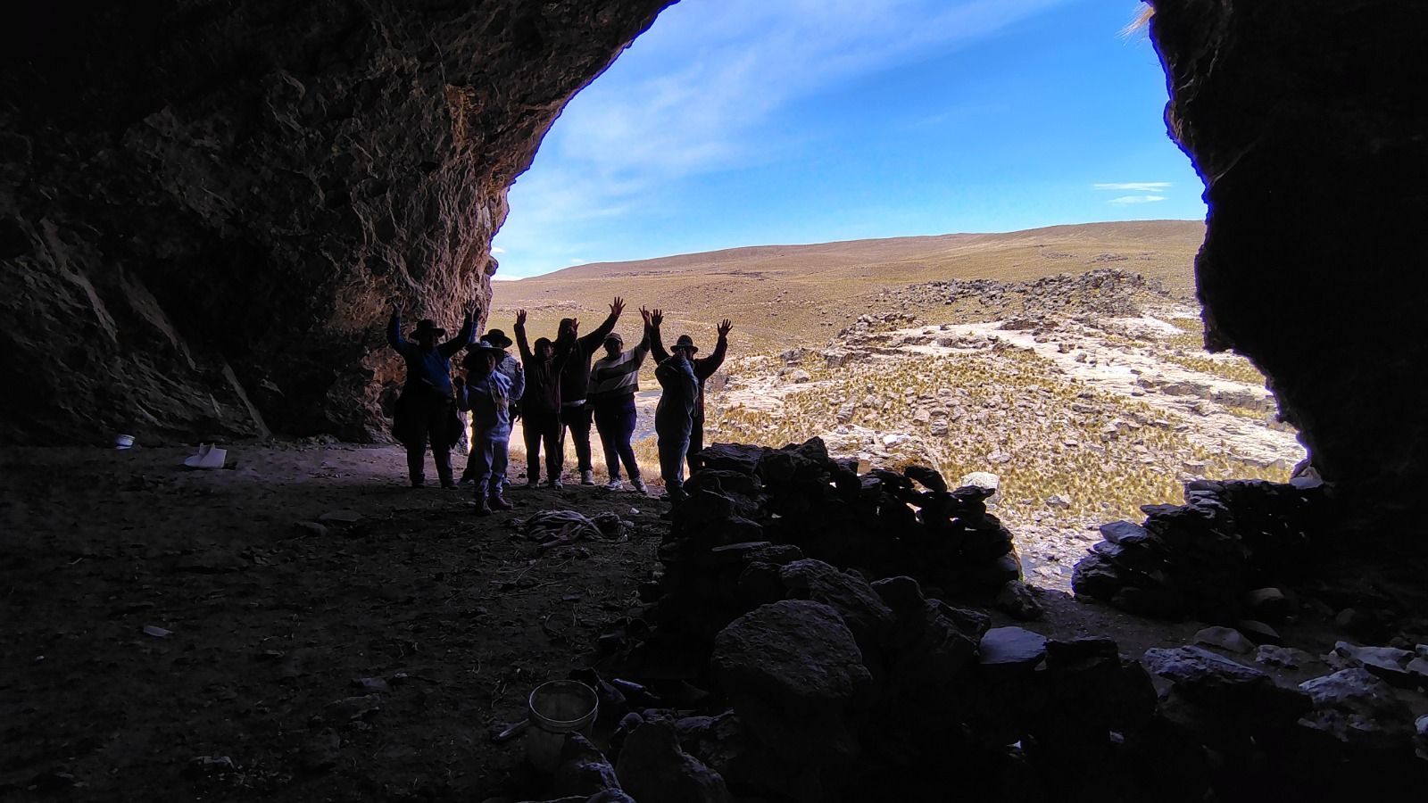 Gente de pie en la entrada de una cueva oscura, contemplando una ladera rocosa iluminada.