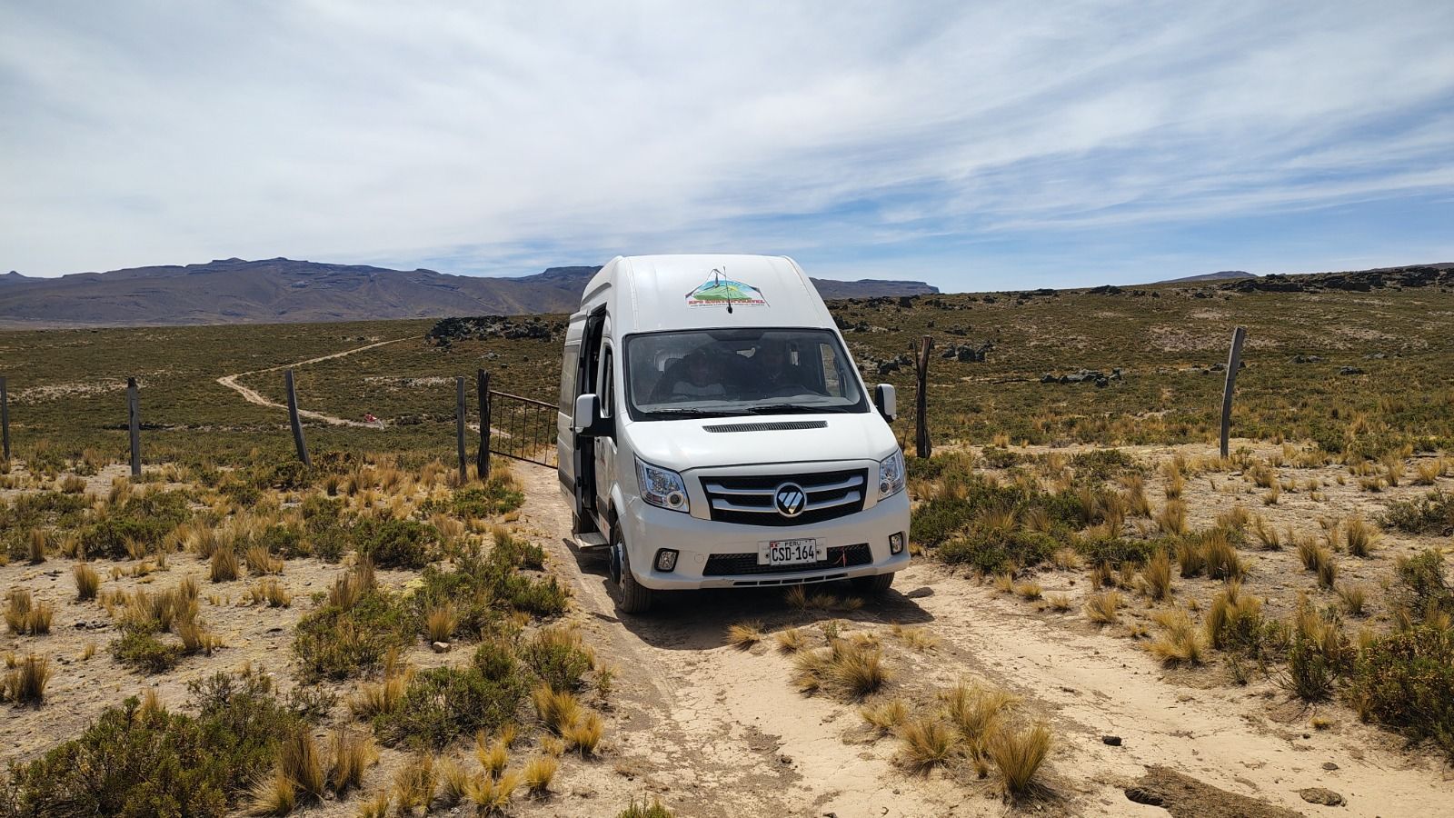 Furgoneta blanca aparcada en un camino de tierra en un paisaje desértico seco y vallado.