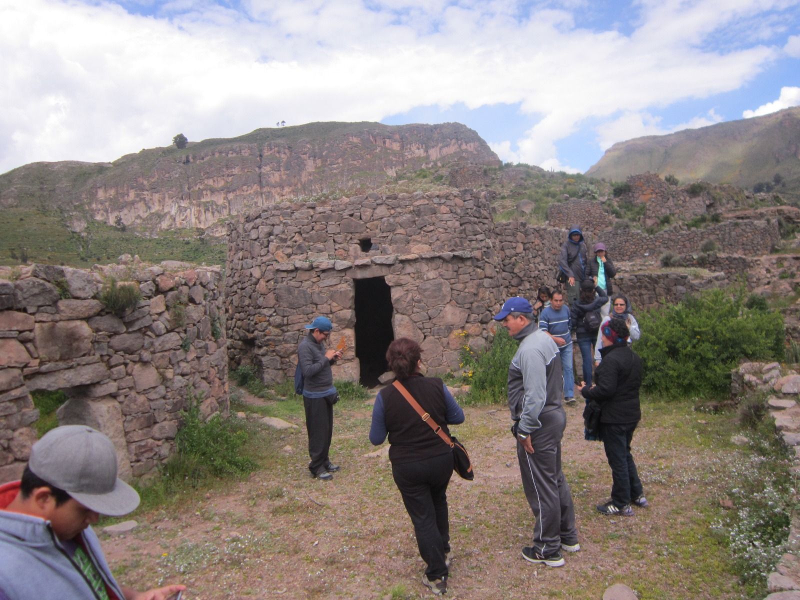 Turistas caminando entre ruinas de piedra en una ladera bajo un cielo nublado.