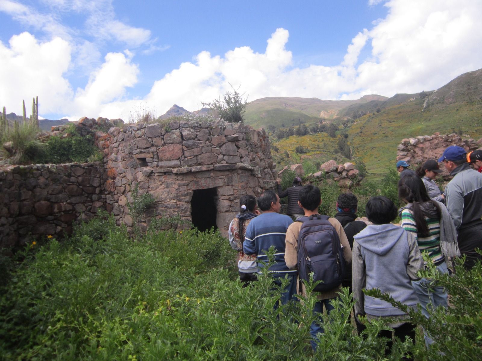 Un grupo de turistas se reúne junto a una ruina de piedra en un valle de montaña verde bajo un cielo nublado.
