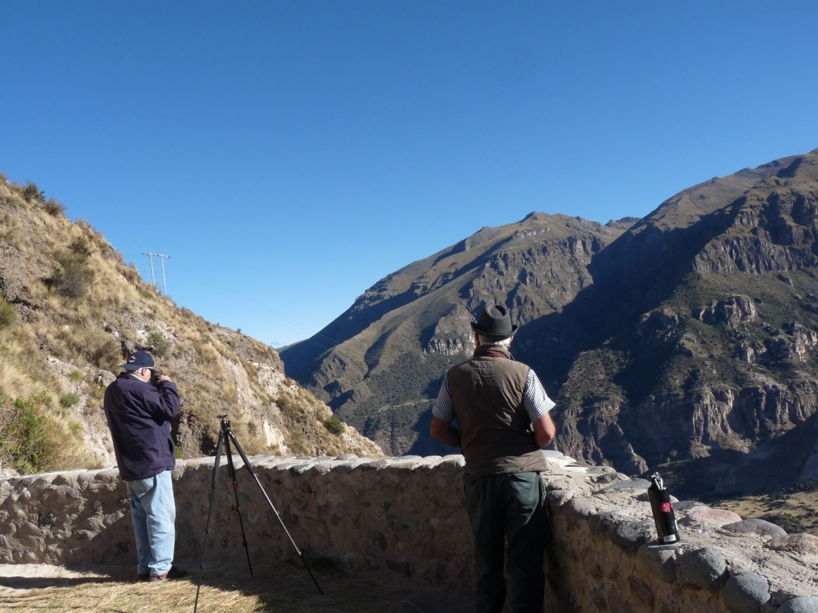 Dos personas fotografiando un valle de montaña desde un mirador de piedra bajo un cielo azul despejado.