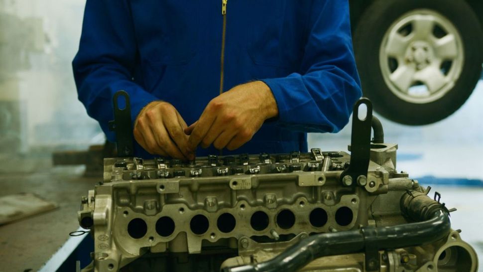 Mecánico con chaqueta azul trabajando en un motor en un taller, con una rueda de coche al fondo.