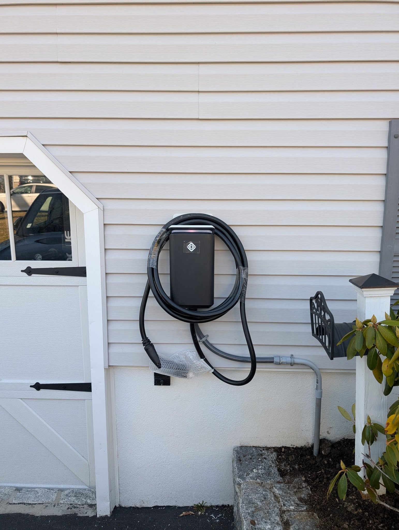 An EV charger with a coiled black cable mounted on light-colored house siding next to a white garage door.
