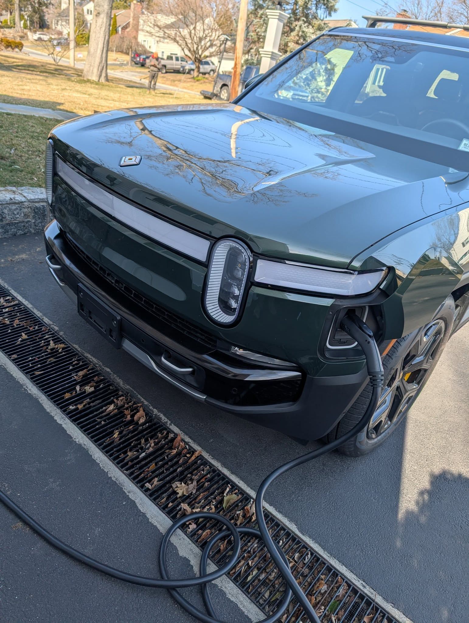 A dark green Rivian electric vehicle parked outdoors and plugged into a charging cable at the front bumper.