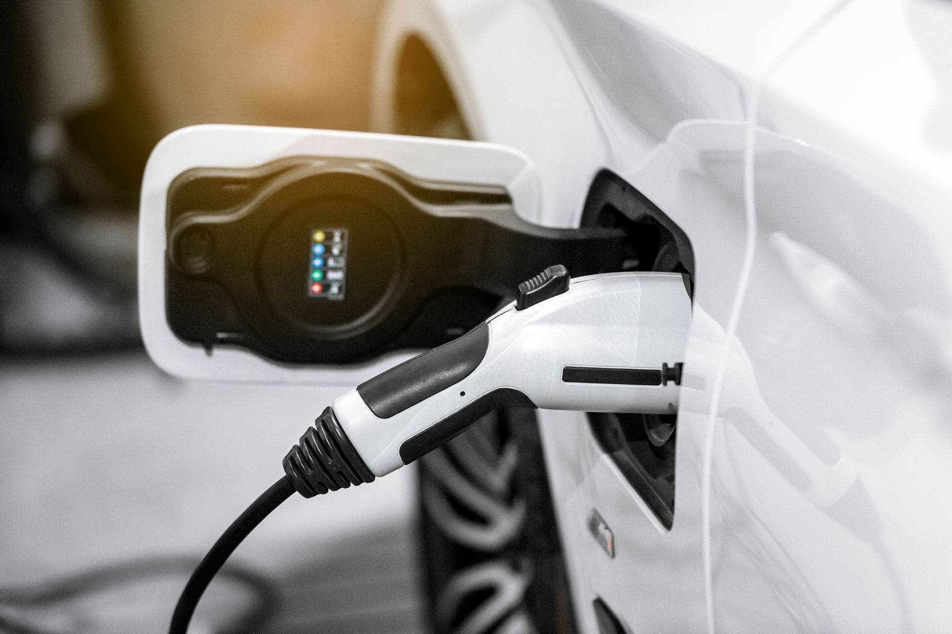 A white electric vehicle plugged into a charging station, with a charging status light visible on the port door.