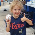 Smiling Tee-ball player in a navy Twins shirt holding a baseball and giving a thumbs-up indoors