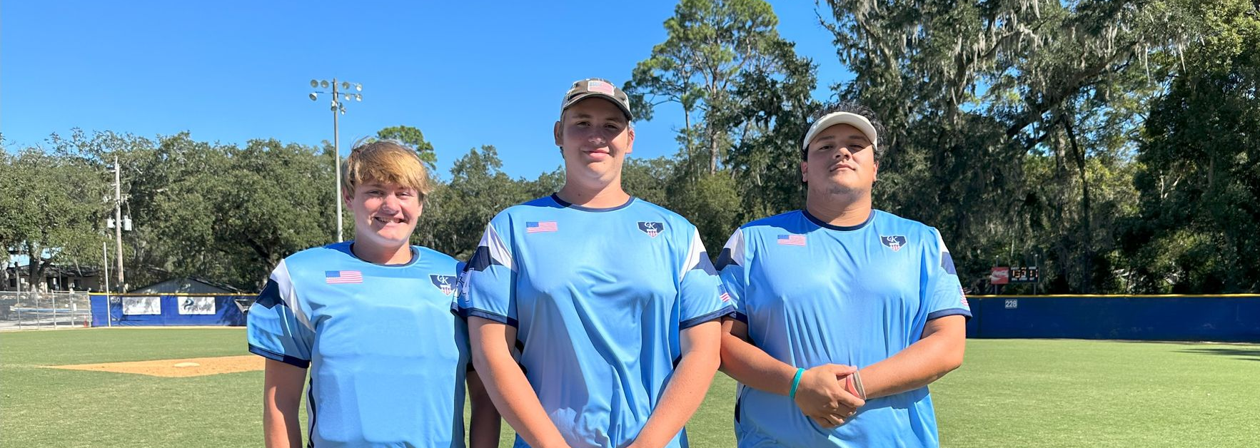 Three people in blue shirts stand on a sunny sports field with trees and a light pole in the background.