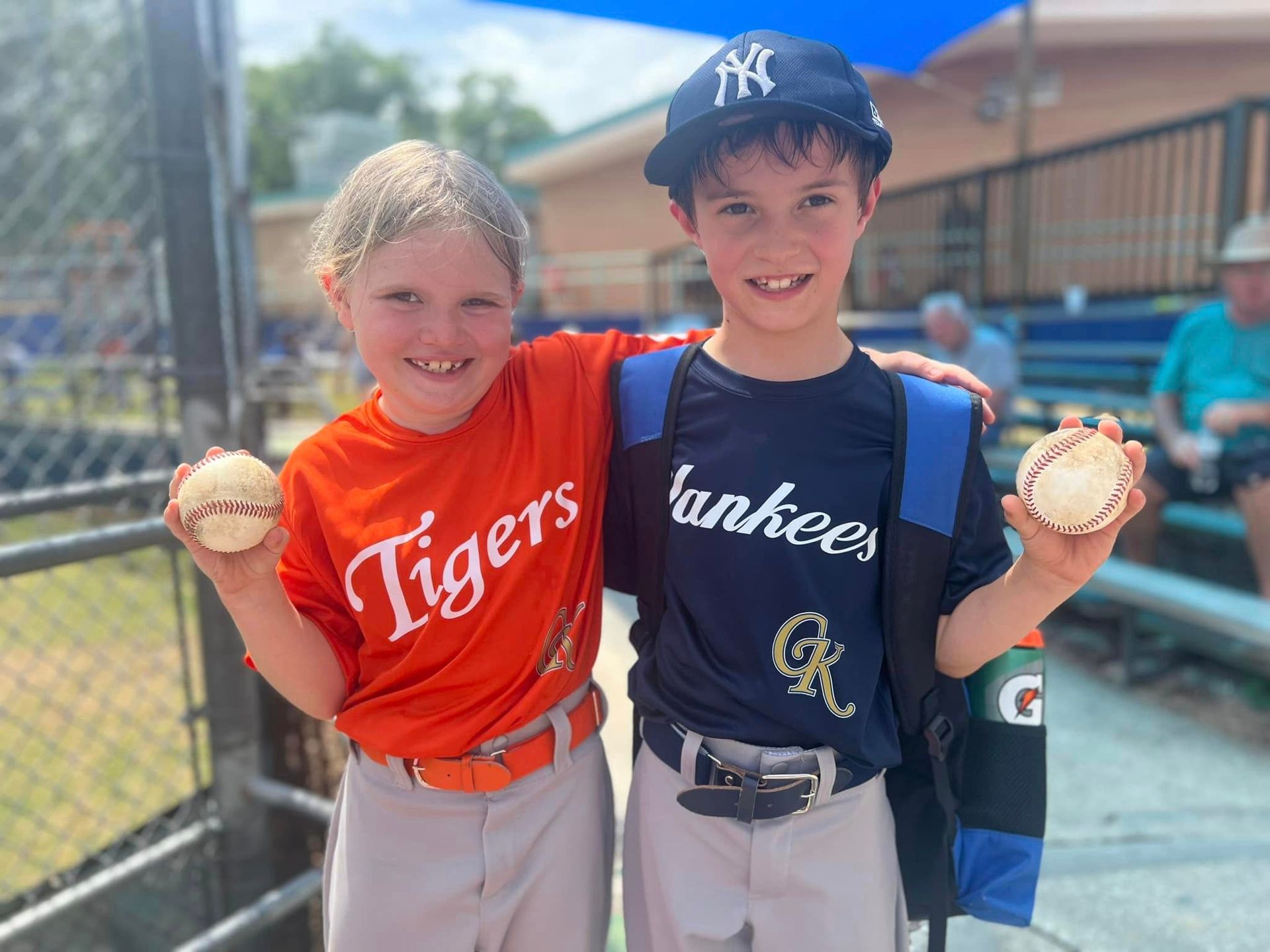 Two youth baseball players smiling and holding balls on a field, wearing Tigers and Yankees jerseys.